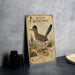 a vintage-style metal sign with illustrations of a roadrunner and desert plants, placed on a countertop next to some black camera lenses.