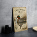 A vintage-style metal sign with an illustration of a green-winged teal duck, placed on a gray countertop next to some black camera lenses.