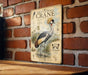 a vintage-style illustration of a grey crowned crane, which is a large wading bird with a distinctive crest on its head. The crane is depicted in a natural setting, surrounded by grass and water. The illustration is mounted on a brick wall, and there is a wooden table below it.