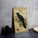 A vintage-style metal sign with a black bird illustration and text, placed on a gray countertop next to some black camera lenses.