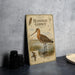 a vintage-style metal sign with illustrations of a bird, a map, and some text, placed on a table next to a stack of black camera lenses.