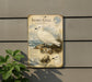 a vintage-style metal plaque with illustrations of two white birds, one standing on a rock and the other sitting on the ground, against a backdrop of a seascape.