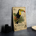 A vintage-style illustration of a toucan, a colorful bird with a large, curved beak, is depicted on a wooden frame. The frame is placed on a gray countertop, with a stack of black camera lenses and a white plate visible in the background.