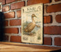 A metal sign with an illustration of a khaki Campbell duck is mounted on a brick wall.