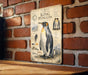 A metal plaque with an illustration of a penguin and various text elements, mounted on a brick wall.
