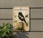 A vintage-style metal sign with illustrations and text about the bird species called "Lark Buntings" is displayed on a wall, with a plant in the foreground.