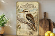 a framed illustration of a laughing kookaburra, a bird native to Australia, perched on a branch. The kookaburra is depicted in a detailed and realistic manner, showcasing its distinctive features such as its large beak, brown and white plumage, and black legs. The frame is placed on a wooden shelf, and there are some potted plants and kitchen utensils visible in the background.