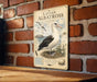 a vintage-style metal plaque with an illustration of a laysan albatross, a large seabird, perched on a nest. The plaque is mounted on a brick wall, and there is a wooden table below it.