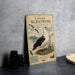 a vintage-style metal plaque with an illustration of a bird, specifically a laysan albatross, standing on a nest. The plaque is placed on a table next to some black camera lenses and a white bowl.