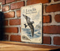 A metal sign with a vintage-style illustration of a bird, likely a seagull, flying over a body of water. The sign is mounted on a brick wall.
