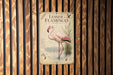 A vintage-style metal sign featuring an illustration of a pink flamingo standing in shallow water, with a wooden fence in the background.