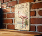 A vintage-style metal sign featuring an illustration of a pink flamingo standing in a body of water, mounted on a brick wall.