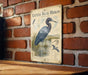 a vintage-style illustration of a blue heron, mounted on a brick wall.