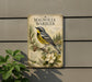 a vintage-style metal sign with a detailed illustration of a bird, specifically a Magnolia Warbler, perched on a branch with a magnolia flower. The sign is mounted on a wall, and there is a plant visible to the left of the sign.