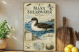 a vintage-style poster or sign about the Manx Shearwater, a bird species, displayed on a shelf with a potted plant and a wooden cutting board.