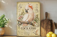 a vintage-style poster of a cockatoo, a type of parrot, displayed on a shelf with various kitchen items such as a wooden cutting board, a lemon, and a potted plant.