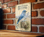 A vintage-style metal sign featuring a blue bird, likely a mountain bluebird, perched on a tree stump against a brick wall background.