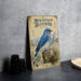 a vintage-style metal sign with a blue bird illustration, placed on a table next to some black camera lenses.