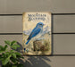 a vintage-style metal sign depicting a blue bird, likely a mountain bluebird, perched on a pine tree stump.