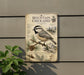 a vintage-style metal sign with illustrations of a mountain chickadee perched on a pine branch, surrounded by pine cones and pine needles. The sign is mounted on a wall and is accompanied by a plant in the foreground.