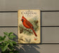 a vintage-style metal sign with a red cardinal perched on a pine branch, surrounded by pine cones and pine needles.