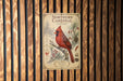 A vintage-style metal sign featuring a red cardinal bird perched on a branch, surrounded by pine cones and berries, mounted on a wooden wall.