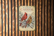 A vintage-style poster of two red cardinals perched on a branch, surrounded by snow and pine needles, is displayed on a wooden wall.