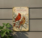 A metal sign with a winter scene of two red cardinals perched on a snow-covered branch, with pine trees and berries in the background.