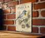 a vintage-style illustration of a bird, specifically the Northern Parula, perched on a branch with pine cones and berries. The bird is depicted in a detailed and colorful manner, showcasing its natural habitat and the flora it feeds on. The illustration is set against a brick wall, adding a rustic and earthy touch to the scene.