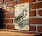 a vintage-style metal sign with an illustration of an osprey, a bird of prey, perched on a branch and holding a fish. The sign is mounted on a brick wall, and there is a wooden table below it.