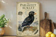 a vintage-style illustration of a parakeet, with a wooden board displaying the title "Parakeetauklet" and additional text. The illustration also includes a detailed illustration of the parakeet in flight and perched on a rock. The background of the illustration depicts a natural landscape with mountains and water. The image is set on a wooden shelf, with a potted plant and a wooden cutting board visible in the background.