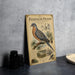 a vintage-style metal sign with an illustration of a passenger pigeon, placed on a table next to some black camera lenses.