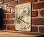 a vintage-style metal plaque with an illustration of a pine siskin bird perched on a branch, surrounded by pine needles and pine cones. The plaque is mounted on a brick wall, and there is a wooden table below it.