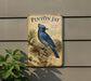 A metal sign with a blue bird perched on a pine tree branch, mounted on a wall.