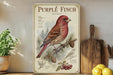 a vintage-style poster of a purple finch, with illustrations of pine cones and berries, displayed on a shelf next to a wooden cutting board and some fruit.