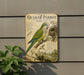 a vintage-style metal sign with a detailed illustration of a Quaker Parrot, including its physical features and habitat. The sign is mounted on a wall, and there is a plant to the left of the sign.