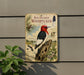 a vintage-style metal sign with illustrations of a red-headed woodpecker, pine cones, and other wildlife elements, mounted on a wall.