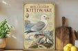 a vintage-style poster of a red-legged Kittiwake bird, with illustrations of the bird, its nest, and its surroundings. The poster is displayed on a wall next to a wooden cutting board and a potted plant.