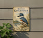 A vintage-style metal sign featuring an illustration of a blue kingfisher bird perched on a tree branch, with a background depicting a river and forest.
