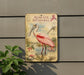 a vintage-style metal sign with illustrations of a pink spoonbill bird, placed on a wall next to a plant.