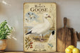 A vintage-style metal plate with an illustration of a white goose, featuring text and a map, is displayed on a shelf next to a potted plant and a wooden cutting board.