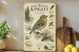 a poster or painting of a Ruby-crowned Kinglet bird, which is a small songbird native to North America. The poster includes detailed illustrations and information about the bird, such as its appearance, habitat, and behavior. The poster is displayed on a wall, with a wooden cutting board and a potted plant visible in the foreground.