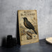 a vintage-style poster of a Rusty Blackbird, a bird known for its black plumage and distinctive black cap. The poster is displayed on a gray wall, and there are some black objects, such as a camera and a bowl, placed nearby.