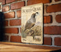 a vintage-style metal sign with a detailed illustration of a quail bird, mounted on a brick wall.