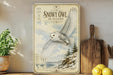 A framed poster of a snowy owl in flight is displayed on a wall, with a wooden cutting board and a potted plant to the side.