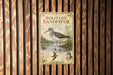 A vintage-style metal sign featuring an illustration of a sandpiper bird standing in a pond, surrounded by plants and trees.