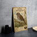 a wooden sign with a picture of a bird, specifically a sparrow, perched on a branch. The sign is placed on a table next to some black camera lenses.