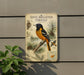 a metal sign with a detailed illustration of a spotted oriole perched on a branch, surrounded by pine cones and other natural elements.