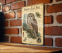 a vintage-style metal sign with an illustration of a spotted owl perched on a branch, mounted on a brick wall.