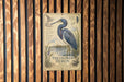 A metal sign with a detailed illustration of a blue heron, mounted on a wooden wall.